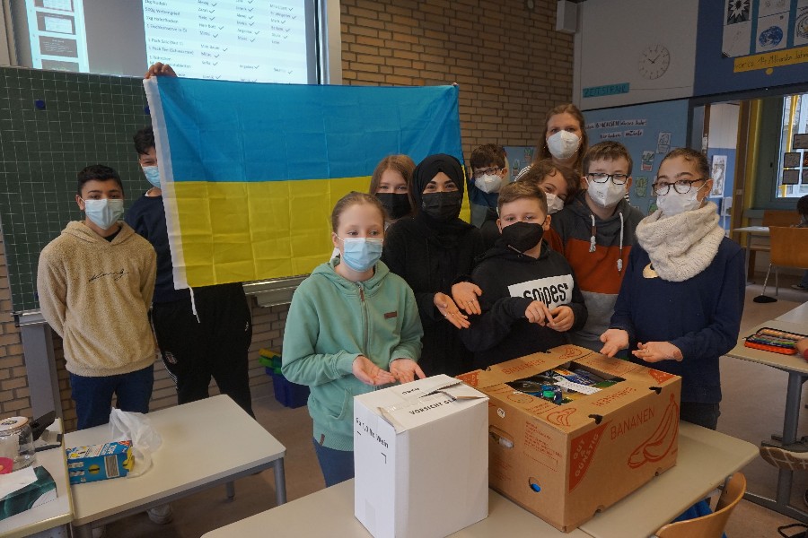 Schülerinnen und Schüler, die vor der blaugelben Ukraine-Flagge stehen und vor ihnen auf dem Tisch stehen die Hilfspakete.