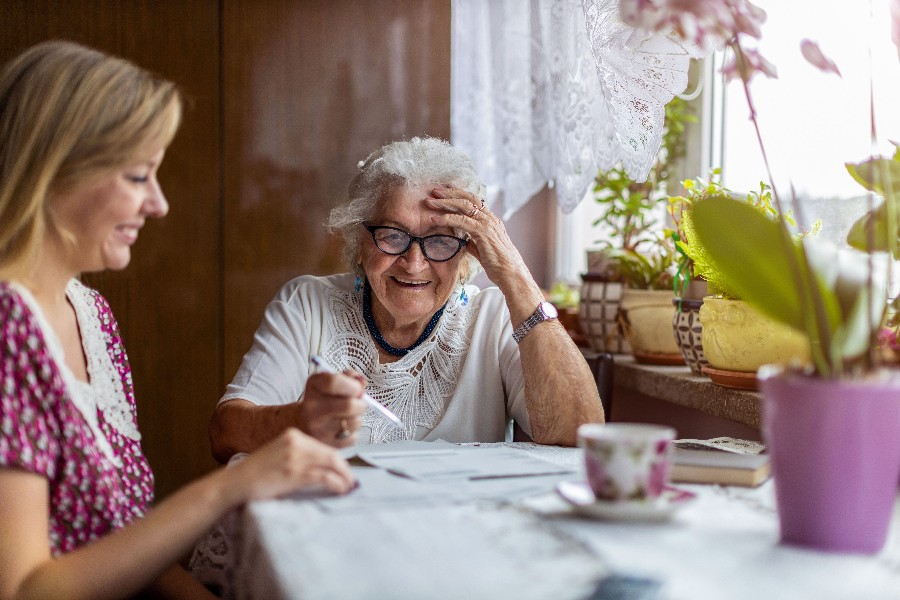 Zu sehen ist eine Seniorin, die mit ihrer Betreuerin an einem Tisch sitzt und Rätsel löst.                  