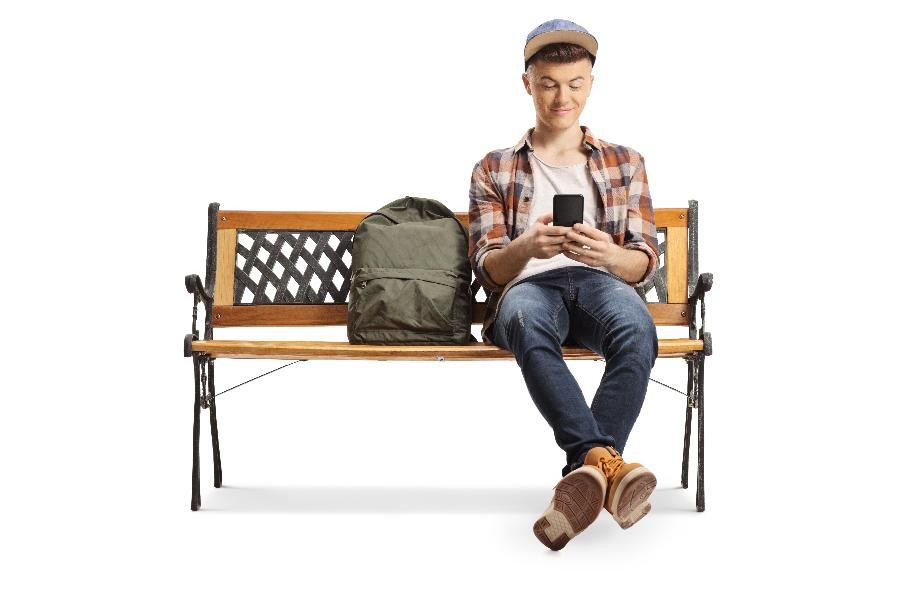 Ein junger Mann sitzt auf einer Holzbank, neben sich einen grünen Rucksack und wartet auf eine Mitfahrgelegenheit.                    