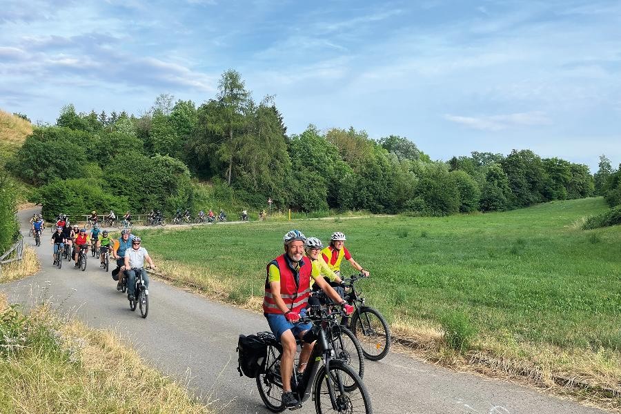 Teilnehmer des Stadtradelns fahren mit ihren Rädern auf einem geteerten Weg entlang einer Wiese.           
