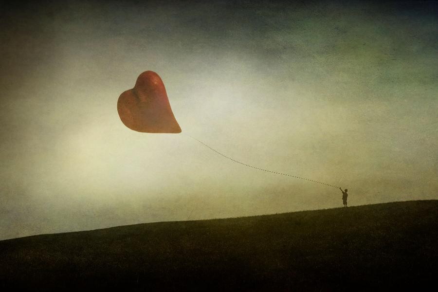 Eine Person steht auf einer dunklen Wiese und hält einen roten Herzluftballon in der Hand, der in den hellen Himmel schwebt.                 