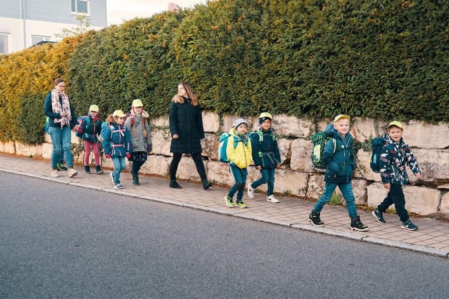 Mehrere Schulkinder laufen mit ihren Begleiterinnen auf dem Gehweg zur Schule.