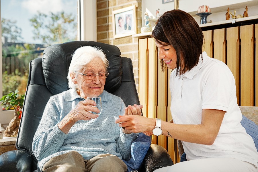 Zu sehen ist eine weibliche Pflegefachkraft, die einer in einem schwarzen Ledersessel sitzenden älteren Dame, die ein Wasserglas in der Hand hält,  ihre Tabletten gibt.              