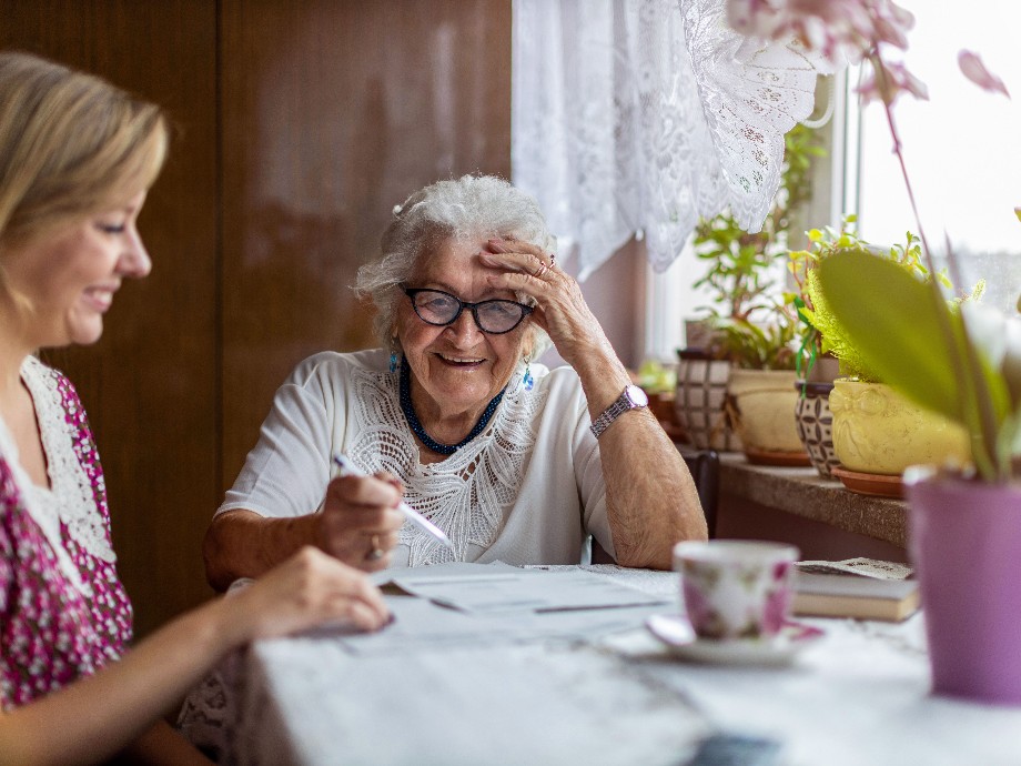 Zu sehen ist eine Seniorin, die mit ihrer Betreuerin an einem Tisch sitzt und Rätsel löst.                  