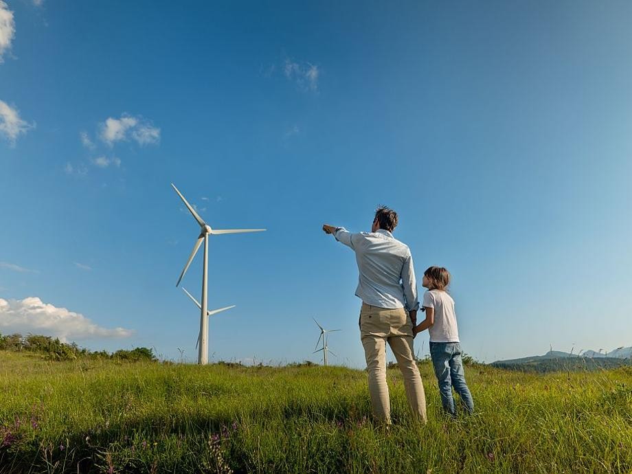 Hier sind unter blauem Himmel und auf einer grünen Wiese Windkrafträder  zu sehen, davor steht ein Mann mit einem Kind an der Hand.