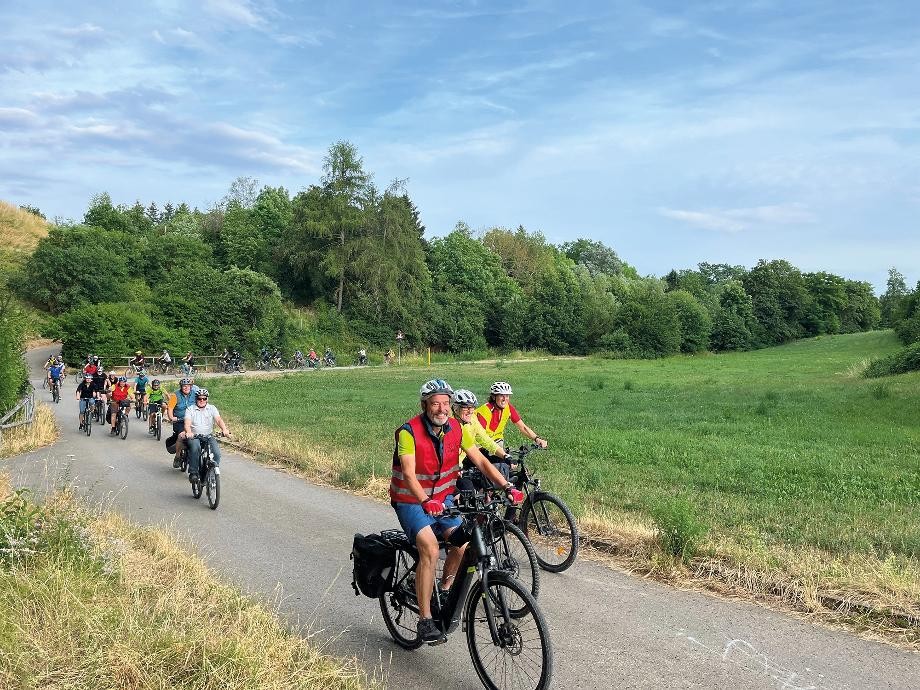 Teilnehmer des Stadtradelns fahren mit ihren Rädern auf einem geteerten Weg entlang einer Wiese.           