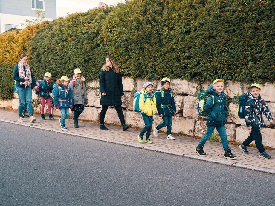 Mehrere Schulkinder laufen mit ihren Begleiterinnen auf dem Gehweg zur Schule.