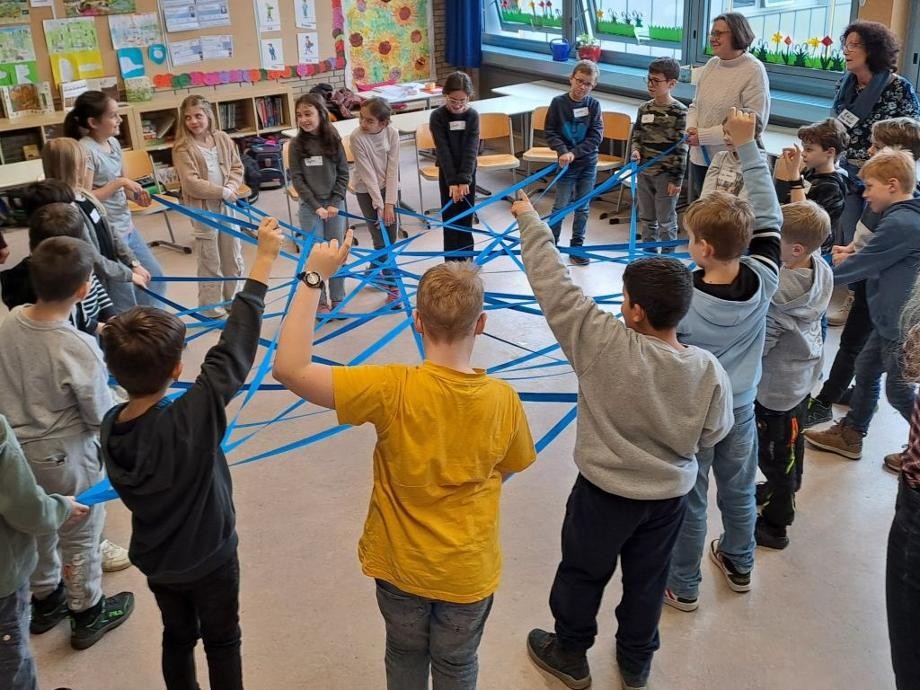 Schülerinnen und Schüler stehen mit ihrem Lehrerinnen im Klassenzimmer im Kreis und halten jeweils über Kreuz blaue Bänder in den Händen.                   