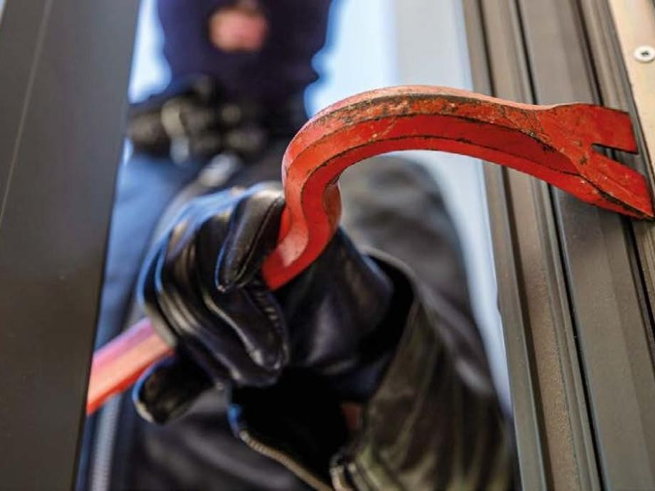 Einbrüche Eine maskierte Person verschafft sich mit einem roten Stemmeisen Zugang zu einer Terrassentür.