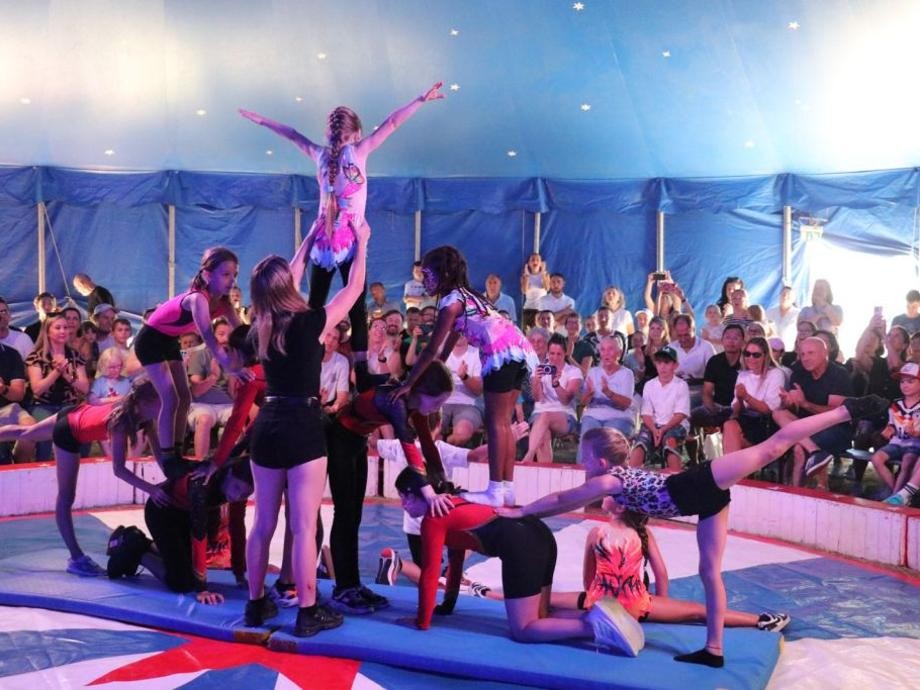Mitmach-Circus Piccolo Schülerinnen führen Akrobatik auf dem Boden des Zirkuszeltes vor und viele Besucherinnen und Besucher schauen zu.