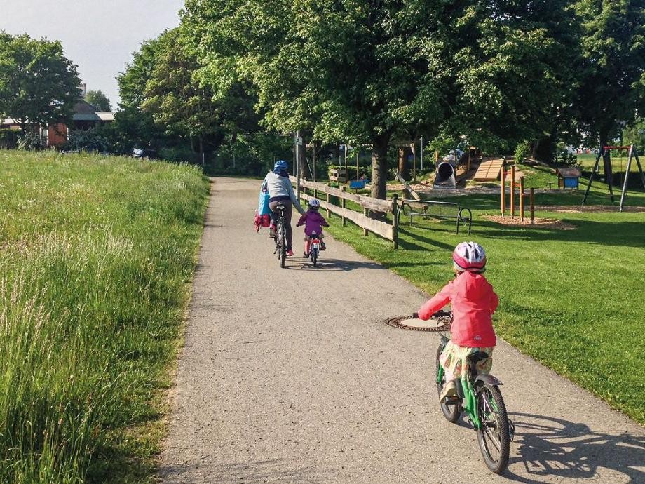 Radfamilie Eine Frau und ihre beiden Kinder fahren mit dem Fahrrad an einem Spielplatz entlang.