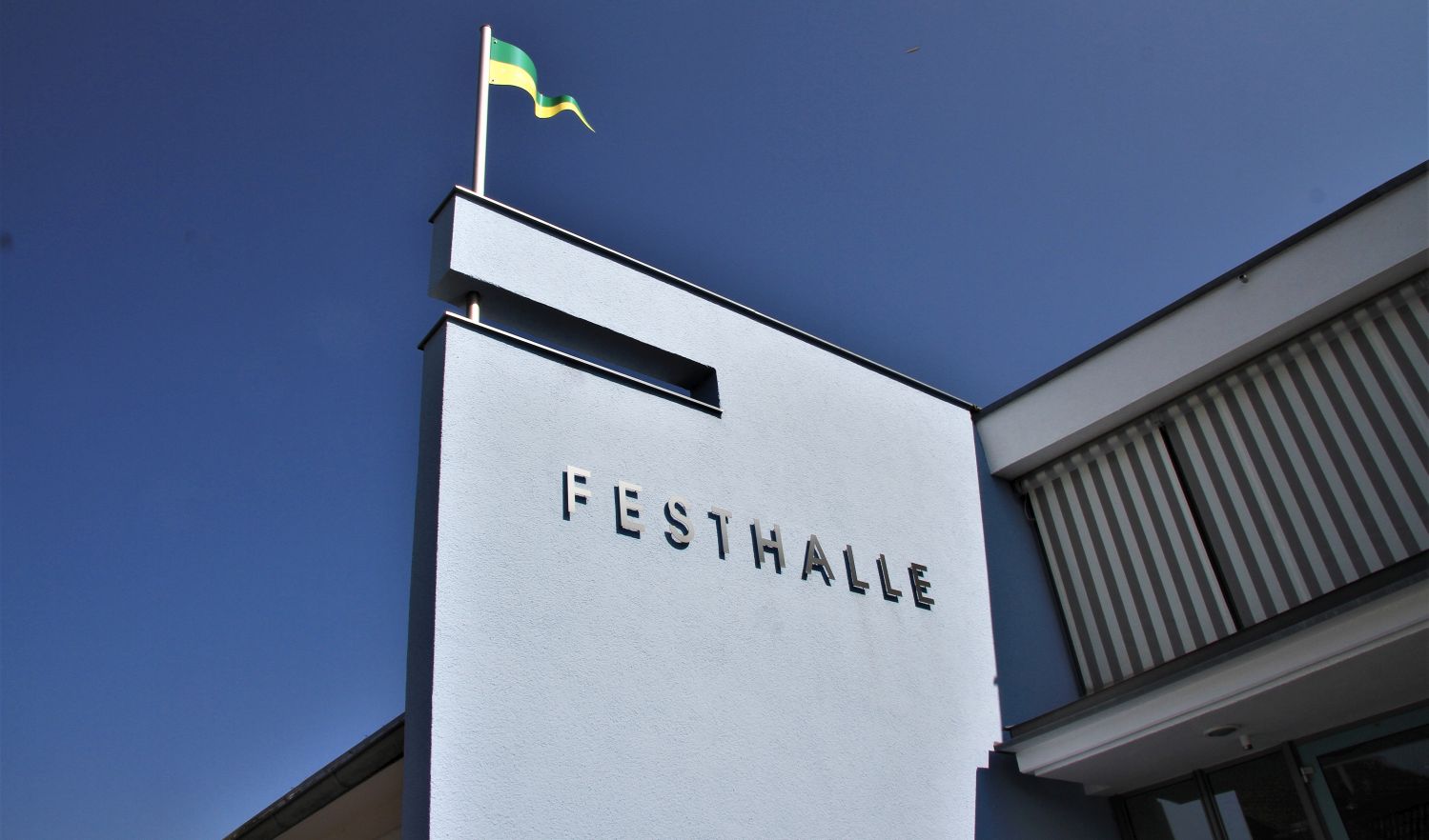 Teilansicht der Fassade der Festhalle Rutesheim Richtung Himmel samt Fahne mit den Stadtfarben grün gelb