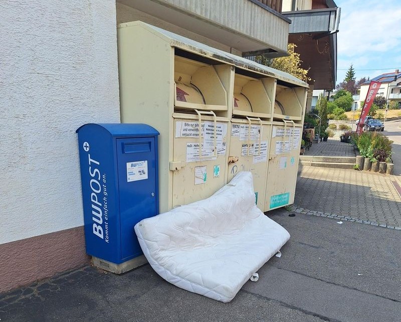 Drei beige Altkleidercontainer und davor liegt eine illegal entsorgte Bettmatratze.